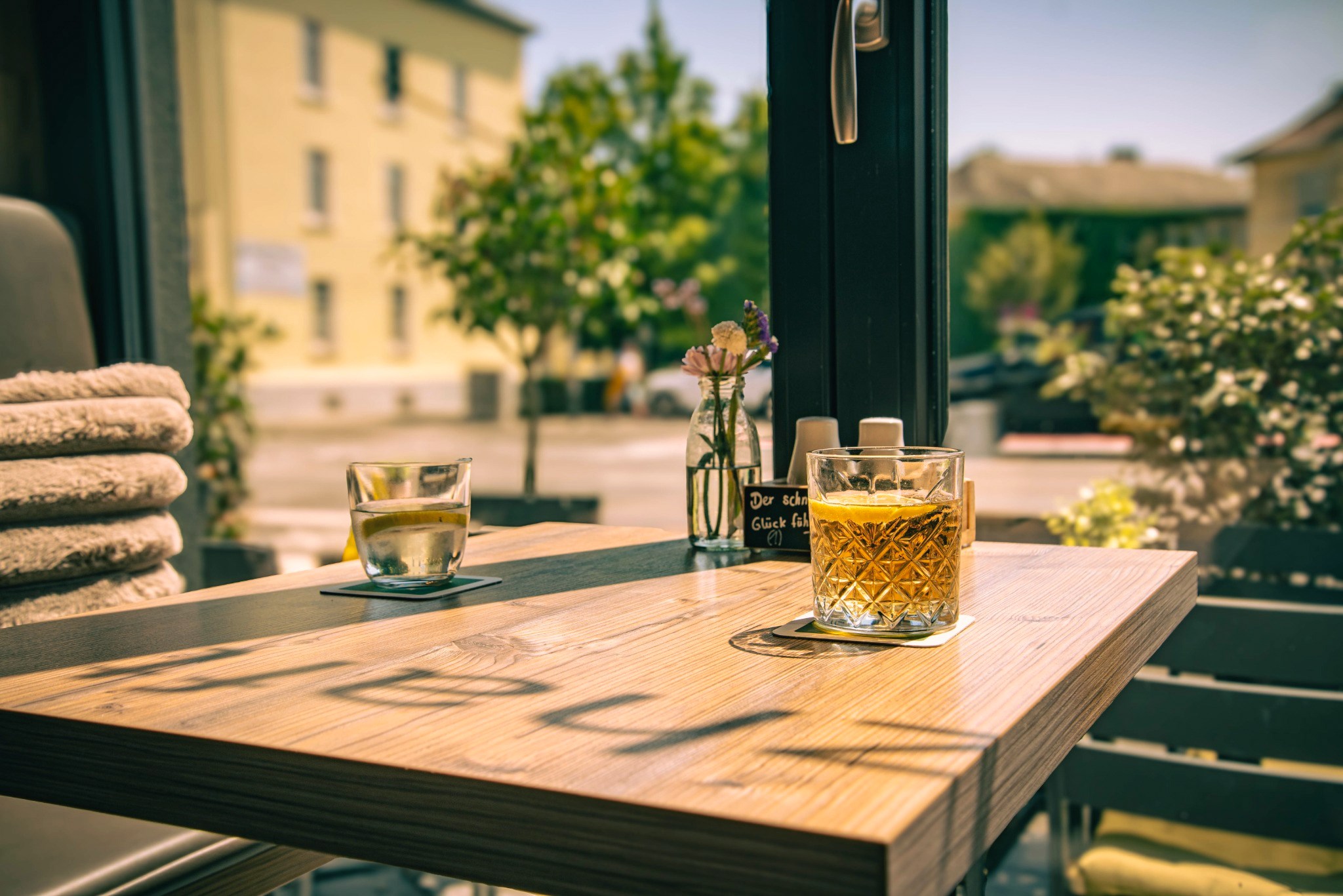 Sonniger Holztisch am Fenster mit zwei Getränken und einer kleinen Blumenvase.